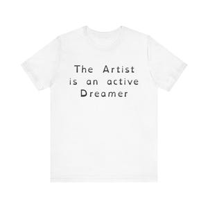 Può includere: Una t-shirt bianca a maniche corte con testo nero stampato sul petto che recita: The Artist is an active Dreamer. Il capo presenta un girocollo e una vestibilità standard, mostrando un design pulito e semplice su un tessuto bianco.
