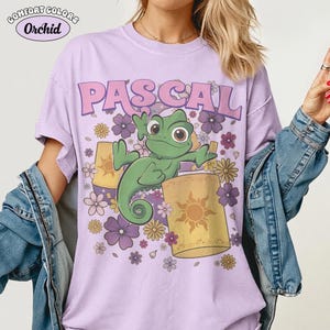 May include: A light purple short-sleeve t-shirt featuring a graphic of a green chameleon named Pascal surrounded by yellow lanterns and purple and yellow flowers. The text PASCAL is printed in large pink letters above the chameleon.