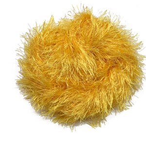 Puede incluir: Una bola redonda de hilo amarillo dorado. El hilo tiene un aspecto texturizado y difuso, con muchos hilos finos que irradian hacia afuera. El material parece suave y podr&iacute;a usarse para varios proyectos de manualidades, como tejer o hacer ganchillo.