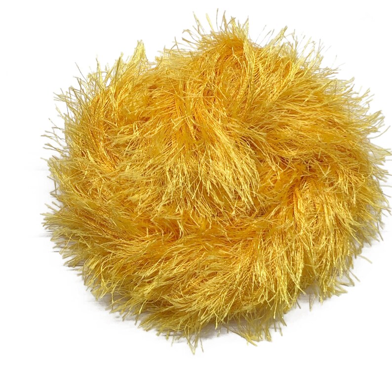 Puede incluir: Una bola redonda de hilo amarillo dorado. El hilo tiene un aspecto texturizado y difuso, con muchos hilos finos que irradian hacia afuera. El material parece suave y podr&iacute;a usarse para varios proyectos de manualidades, como tejer o hacer ganchillo.