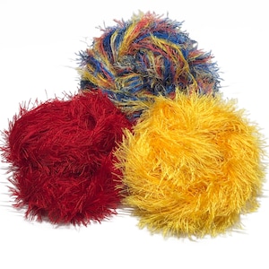 Puede incluir: Tres ovillos de lana esponjosa en colores vibrantes: rojo, amarillo y una mezcla de azul, amarillo y rojo. La lana tiene un aspecto texturizado y difuso, ideal para proyectos de manualidades. Los ovillos est&aacute;n sobre un fondo blanco.