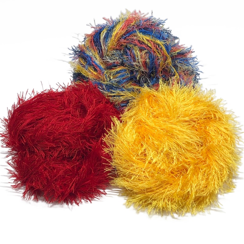 Puede incluir: Tres ovillos de lana esponjosa en colores vibrantes: rojo, amarillo y una mezcla de azul, amarillo y rojo. La lana tiene un aspecto texturizado y difuso, ideal para proyectos de manualidades. Los ovillos est&aacute;n sobre un fondo blanco.