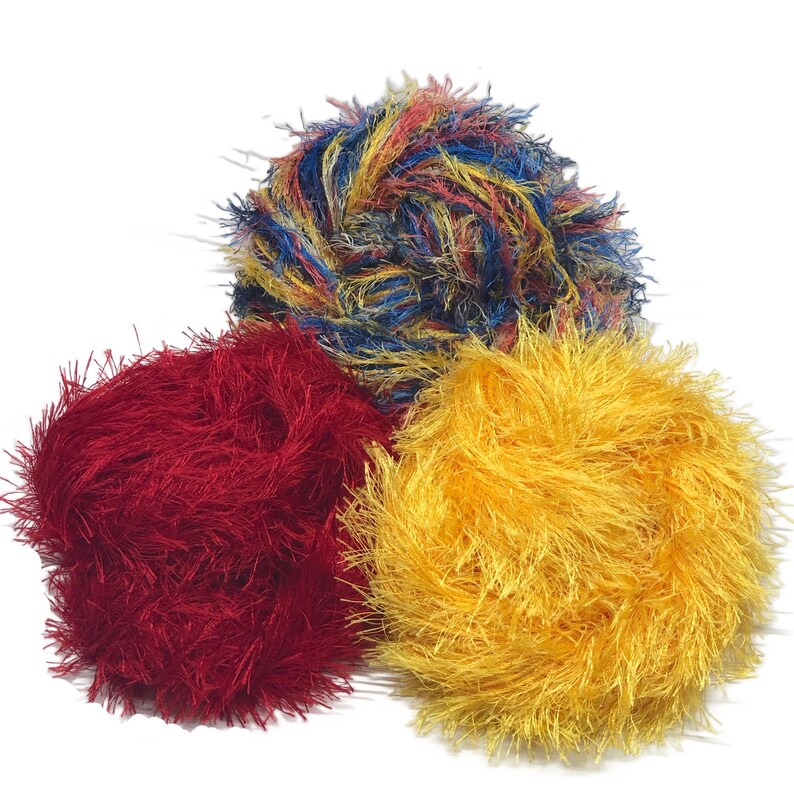 Puede incluir: Tres bolas de hilo esponjosas en rojo, amarillo y una mezcla de azul, amarillo y rojo. El hilo tiene una textura borrosa, y las bolas est&aacute;n dispuestas en forma triangular sobre un fondo blanco. Ideal para manualidades.