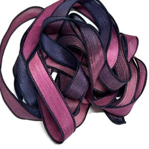 May include: A close-up of a spool of ombre ribbon with a dark blue and pink color gradient. The ribbon has a black edge and is made of a soft, silky material.