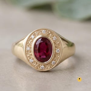 Op de afbeelding: Een gouden ring met een centrale ovaal geslepen dieprode edelsteen, omringd door een decoratieve rand met gegraveerde bladmotieven en kleine ronde witte diamanten op een gepolijste gouden band met een glad, afgerond ontwerp.