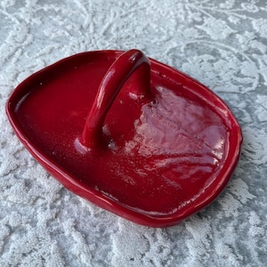 Può includere: Un vassoio in ceramica rosso brillante e lucido, di forma ovale, con un unico manico ad arco che si estende dal centro. La superficie presenta una finitura liscia e riflettente con bordi leggermente rialzati e irregolari.