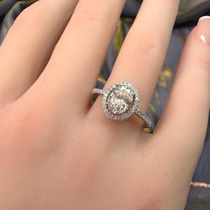 May include: A silver-toned ring featuring a large, oval-cut center stone surrounded by a halo of smaller, round-cut stones. The band is slender and also encrusted with tiny, sparkling stones along the visible upper portion of the ring.
