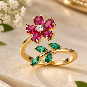 May include: A gold ring featuring a flower motif with five pear-cut deep pink stones surrounding a central clear round stone, complemented by a branch-like design adorned with four marquise-cut green stones along the band.