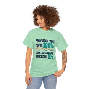 May include: A light green short-sleeve t-shirt featuring dark blue text that reads: FROM 100 FEET AWAY, YOU'RE 100%. SADLY, EVERY FOOT CLOSER KNOCKS OFF 1%. The design includes a horizontal line with small orange footprints above it.