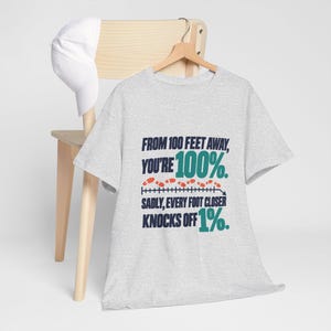 May include: A light gray short-sleeve t-shirt featuring dark blue and teal text that reads: FROM 100 FEET AWAY, YOU'RE 100%. SADLY, EVERY FOOT CLOSER KNOCKS OFF 1%. A graphic of small orange footprints follows a line between the text.