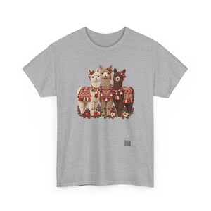 May include: A heather gray t-shirt featuring a graphic of three alpacas in white, tan, and dark brown. Each alpaca wears a colorful patterned blanket with tassels and floral headpieces. The alpacas stand among small flowers at the base.