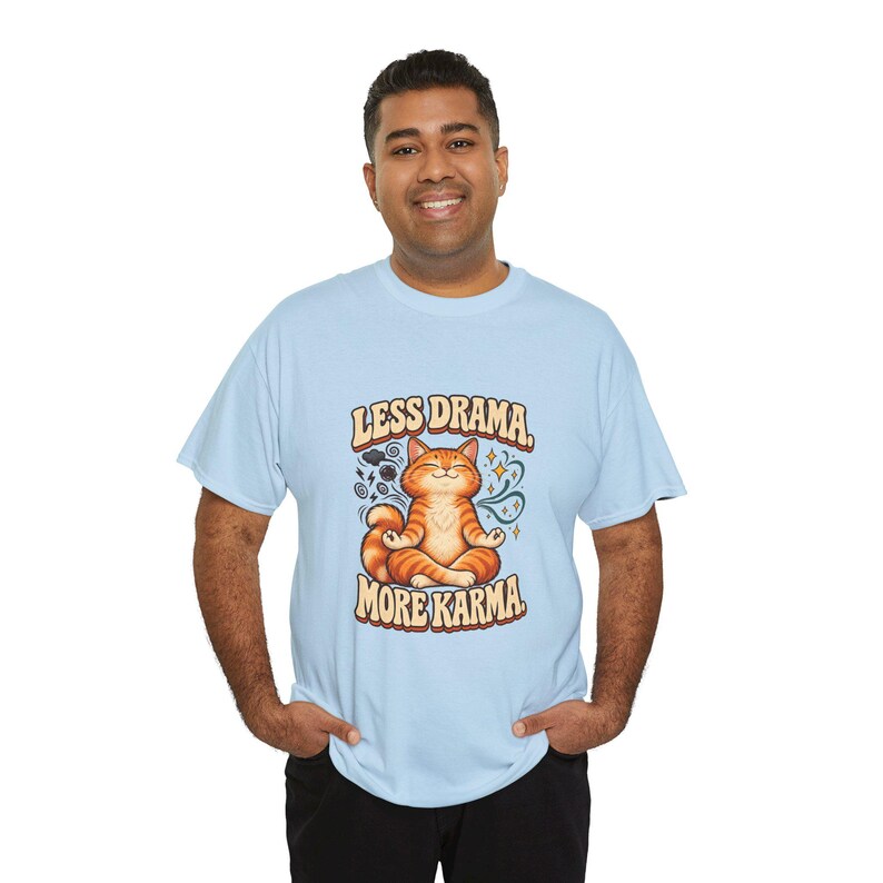May include: A light blue short-sleeve t-shirt featuring a graphic of an orange tabby cat sitting in a lotus position. The text above the cat reads LESS DRAMA and the text below reads MORE KARMA. The cat is surrounded by small stars and swirls.