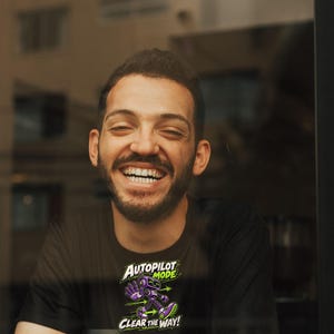 May include: A black t-shirt featuring a graphic of a purple robot with green accents. The text above the robot reads AUTOPILOT MODE and the text below reads CLEAR THE WAY! The design includes green arrows pointing in opposite directions.