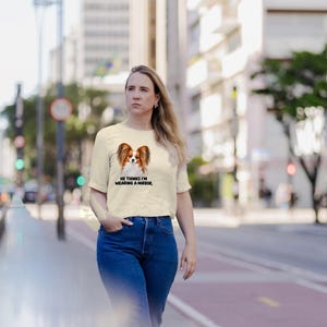 K&ouml;nnte beinhalten: Ein cremefarbenes Kurzarm-T-Shirt mit einer Grafik eines Papillon-Hundes und dem schwarzen Text "HE THINKS I'M WEARING A MIRROR." Das T-Shirt hat einen Rundhalsausschnitt und einen geraden Saum, kombiniert mit einer blauen Jeans.