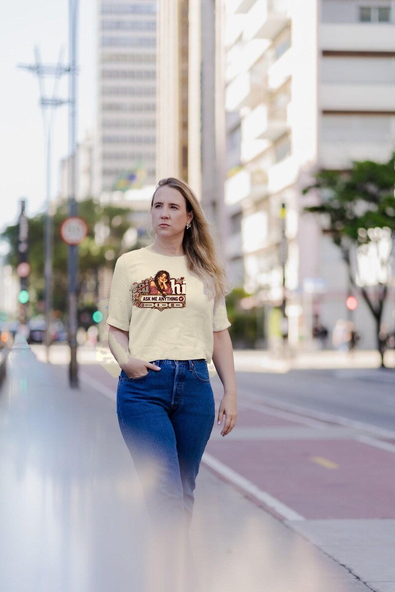 May include: A cream-colored short-sleeve t-shirt featuring a graphic print on the chest. The design includes an illustration of a person with long dark hair, the text "hi" and "ASK ME ANYTHING," and decorative floral elements in shades of red and brown.