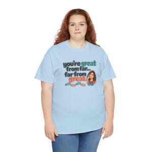 May include: A light blue short-sleeve t-shirt featuring a graphic print with the text "you're great from far... far from great." The design includes a cartoon illustration of a person looking through binoculars and small footprints.