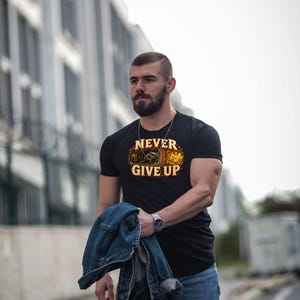 Puede incluir: Una camiseta negra de manga corta con un estampado gr&aacute;fico de un minero trabajando en una cueva y el texto NEVER GIVE UP en letras amarillas. El dise&ntilde;o incluye tonos naranjas y marrones con una escena de cristales brillantes.