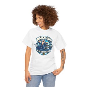 May include: A white short-sleeve t-shirt featuring a circular graphic of a pilot and a robot in a cockpit. The text reads WATCH OUT! I'M ON AUTOPILOT NOW. The design uses blue, white, and yellow colors with wings on the sides of the circle.