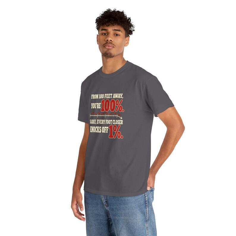 May include: A dark gray short-sleeve t-shirt featuring the text: FROM 100 FEET AWAY, YOU'RE 100%. SADLY, EVERY FOOT CLOSER KNOCKS OFF 1%. The text includes a graphic line with a downward arrow and uses red and white lettering.