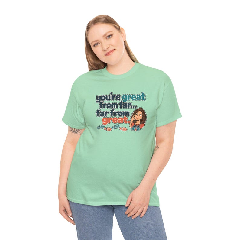May include: A light green short-sleeve t-shirt featuring the text "you're great from far... far from great." The graphic includes a cartoon illustration of a person holding a game controller and three small footprints in blue and red.