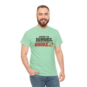 May include: A light green short-sleeve t-shirt featuring the text "HARD TO IGNORE. ESPECIALLY WHEN I SNORE." in dark green and red lettering, accompanied by a small cartoon illustration of a person sleeping with a speech bubble.