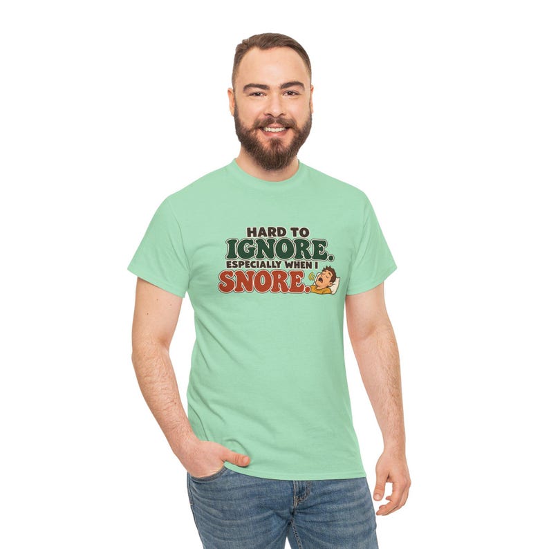 May include: A light green short-sleeve t-shirt featuring the text "HARD TO IGNORE. ESPECIALLY WHEN I SNORE." in dark green and red lettering, accompanied by a small cartoon illustration of a person sleeping with a speech bubble.
