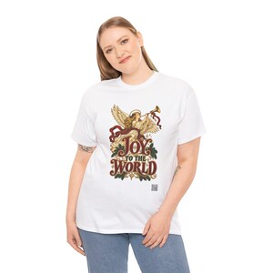 May include: A white short-sleeve t-shirt featuring a graphic of an angel playing a trumpet with red ribbons and holly leaves. The text reads Joy to the World in a decorative font. The design includes small star accents and a small QR code.