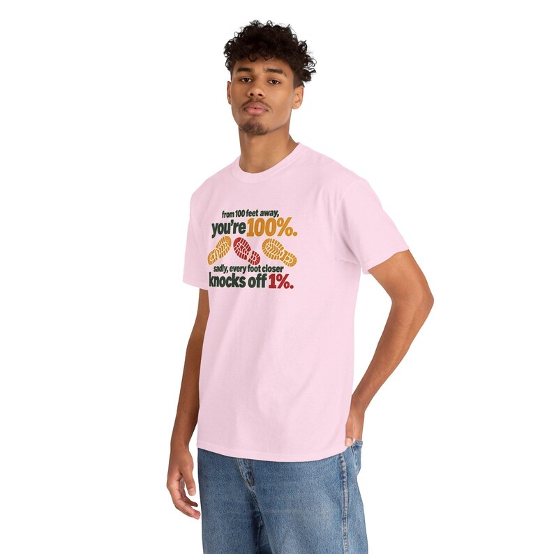 May include: A light pink short-sleeve t-shirt featuring the black and red text: from 100 feet away, you're 100%. sadly, every foot closer knocks off 1%. The design includes three small, colorful shoe print graphics in yellow and red.