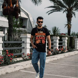 May include: A black short-sleeve t-shirt featuring the text I AM MEDIUM LOOKING FOR RARE with cartoon steak illustrations, paired with blue denim jeans and white athletic sneakers. The shirt displays a graphic of two steaks with faces.
