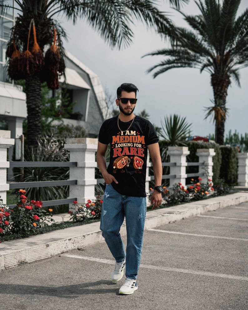 May include: A black short-sleeve t-shirt featuring the text I AM MEDIUM LOOKING FOR RARE with cartoon steak illustrations, paired with blue denim jeans and white athletic sneakers. The shirt displays a graphic of two steaks with faces.