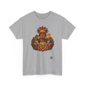 May include: A heather gray t-shirt featuring a central graphic of a person wearing an elaborate feathered headdress and gold jewelry. The design includes mountain silhouettes, a sun, and geometric patterns in red, gold, and green.
