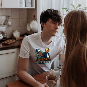 May include: A white short-sleeved t-shirt featuring a graphic illustration of a person with a hand raised in a stop gesture, a cigarette, and blue smoke clouds. The text printed on the shirt reads, "SMOKE THERE, I BREATHE HERE."