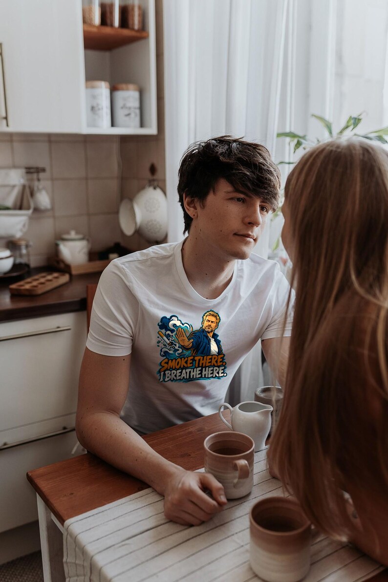 May include: A white short-sleeved t-shirt featuring a graphic illustration of a person with a hand raised in a stop gesture, a cigarette, and blue smoke clouds. The text printed on the shirt reads, "SMOKE THERE, I BREATHE HERE."