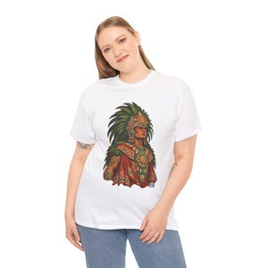 May include: A white short-sleeved t-shirt featuring a graphic print of an Aztec warrior wearing a large green feathered headdress, a gold sun-disk chest plate, and ornate red and green patterned clothing with gold accents.