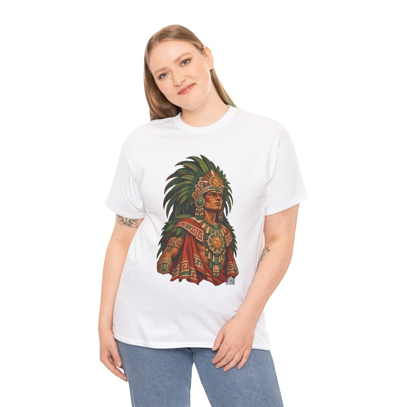 May include: A white short-sleeved t-shirt featuring a graphic print of an Aztec warrior wearing a large green feathered headdress, a gold sun-disk chest plate, and ornate red and green patterned clothing with gold accents.