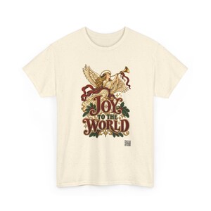 May include: A cream-colored short-sleeve t-shirt with a graphic print of an angel playing a trumpet. The text reads "JOY TO THE WORLD" in a decorative font, surrounded by holly leaves, red ribbons, and small stars on the front.