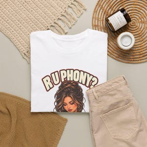 May include: A white t-shirt featuring a graphic of a person with voluminous brown hair and the text RU PHONY? in a curved, bold font. The design includes a person with hoop earrings and a detailed hairstyle centered on the chest area.