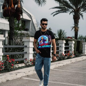 Może przedstawiać: Czarny t-shirt z kr&oacute;tkim rękawem z nadrukiem graficznym przedstawiającym osobę z wyciągniętą ręką, otoczoną niebieską sferą energii i fioletowym dymem, z napisem NOT IN MY AIRSPACE niebieskimi i r&oacute;żowymi literami, zestawiony z niebieskimi dżinsami.