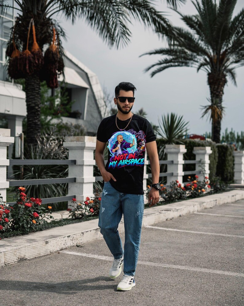 Może przedstawiać: Czarny t-shirt z kr&oacute;tkim rękawem z nadrukiem graficznym przedstawiającym osobę z wyciągniętą ręką, otoczoną niebieską sferą energii i fioletowym dymem, z napisem NOT IN MY AIRSPACE niebieskimi i r&oacute;żowymi literami, zestawiony z niebieskimi dżinsami.