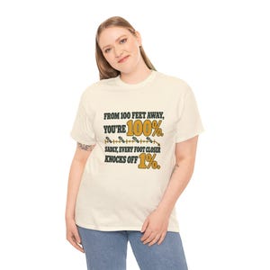 May include: A cream-colored short-sleeve t-shirt with black and gold text that reads: FROM 100 FEET AWAY, YOU'RE 100%. SADLY, EVERY FOOT CLOSER KNOCKS OFF 1%. The design includes a graphic of a measuring line with small footprints.