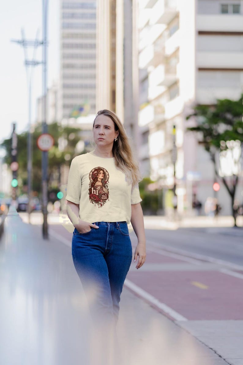 K&ouml;nnte beinhalten: Ein cremefarbenes Kurzarm-T-Shirt mit einem zentralen Grafikdruck einer Frau mit langem braunem Haar, umgeben von verzierten Blumenmustern und roten Rosen, mit dem kleingeschriebenen Text "hi ai" in einer dunkelbraunen Serifenschrift.