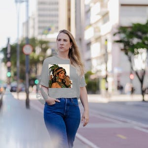 Puede incluir: Una camiseta gris claro de manga corta con un estampado gr&aacute;fico rectangular de una persona con un tocado de plumas verdes, joyas doradas y pendientes verdes, combinada con unos vaqueros de color azul oscuro.