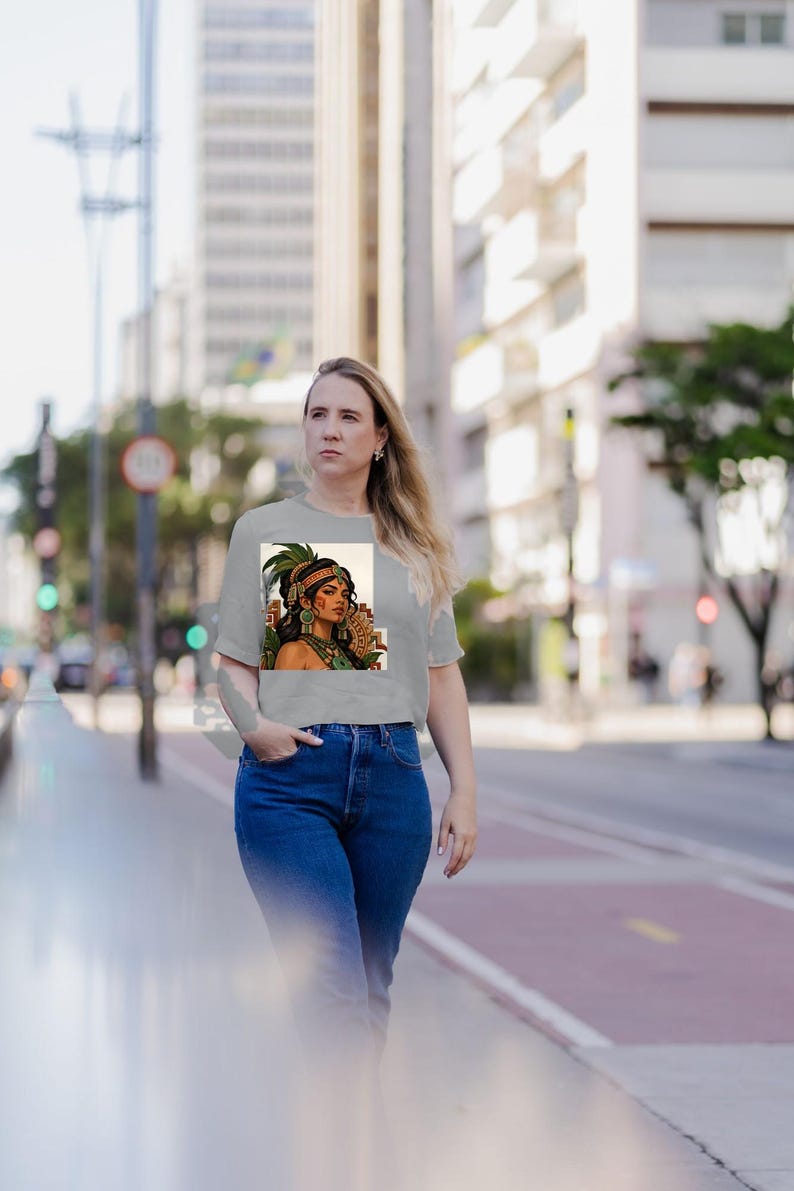 Puede incluir: Una camiseta gris claro de manga corta con un estampado gr&aacute;fico rectangular de una persona con un tocado de plumas verdes, joyas doradas y pendientes verdes, combinada con unos vaqueros de color azul oscuro.