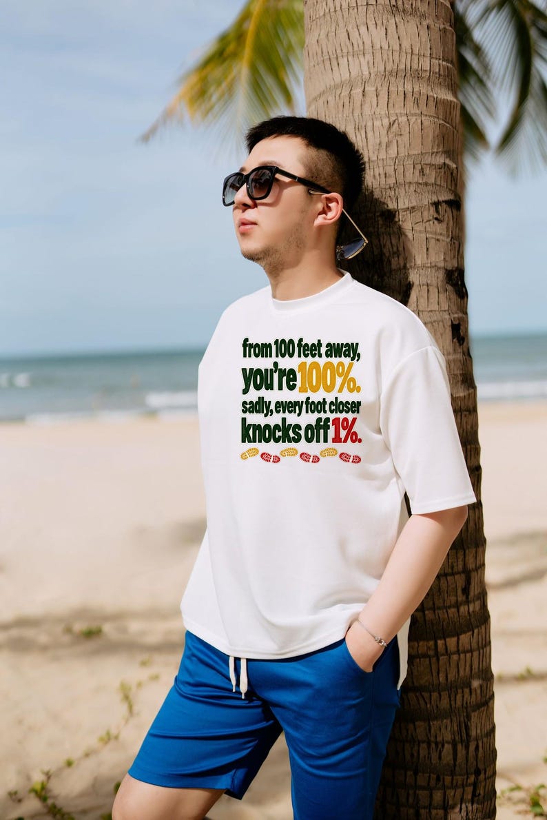 May include: A white short-sleeved t-shirt featuring the dark green and red text: from 100 feet away, you're 100%. sadly, every foot closer knocks off 1%. The design includes a row of small red and yellow shoe print icons below the text.