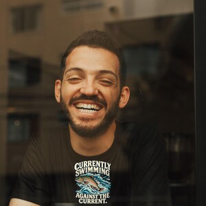 May include: A black short-sleeved t-shirt featuring a graphic of a fish jumping out of blue water waves with the text "CURRENTLY SWIMMING AGAINST THE CURRENT." The shirt is worn by a person sitting behind a white coffee cup with a black lid.