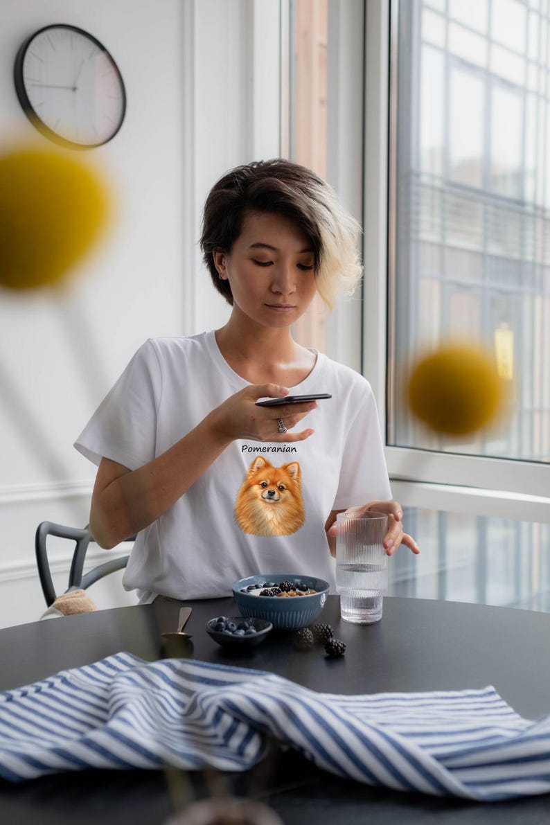 May include: A white short-sleeved t-shirt featuring a graphic of a brown Pomeranian dog on the chest with the text "Pomeranian" printed in black lettering above the dog's head.
