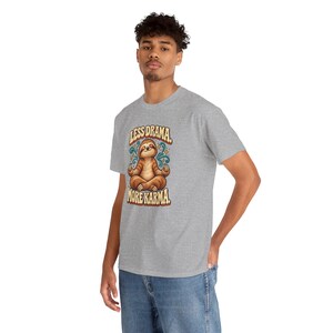 May include: A heather gray short-sleeve t-shirt featuring a graphic of a brown sloth sitting in a lotus position. The text above and below the sloth reads Less Drama. More Karma. The design includes teal swirl accents and small yellow stars.