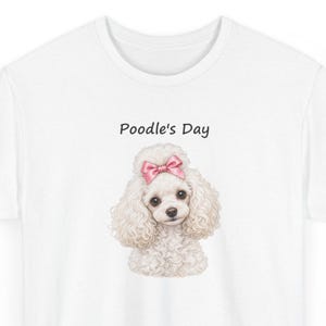 May include: A white t-shirt featuring a graphic of a fluffy white poodle with a pink bow on its head. Above the poodle, the text Poodle's Day is printed in a simple black font. The design is centered on the chest of the shirt.
