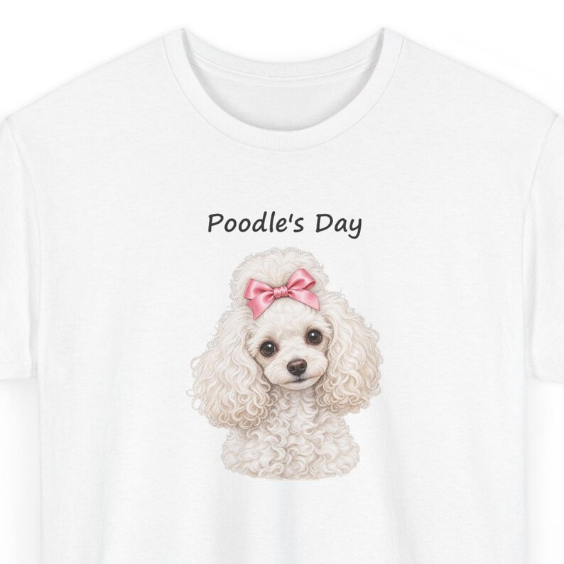 May include: A white t-shirt featuring a graphic of a fluffy white poodle with a pink bow on its head. Above the poodle, the text Poodle's Day is printed in a simple black font. The design is centered on the chest of the shirt.