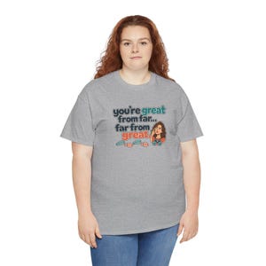 May include: A heather gray short-sleeve t-shirt featuring the text "you're great from far... far from great." with a cartoon illustration of a person looking through binoculars and small colorful footprint icons below the text.